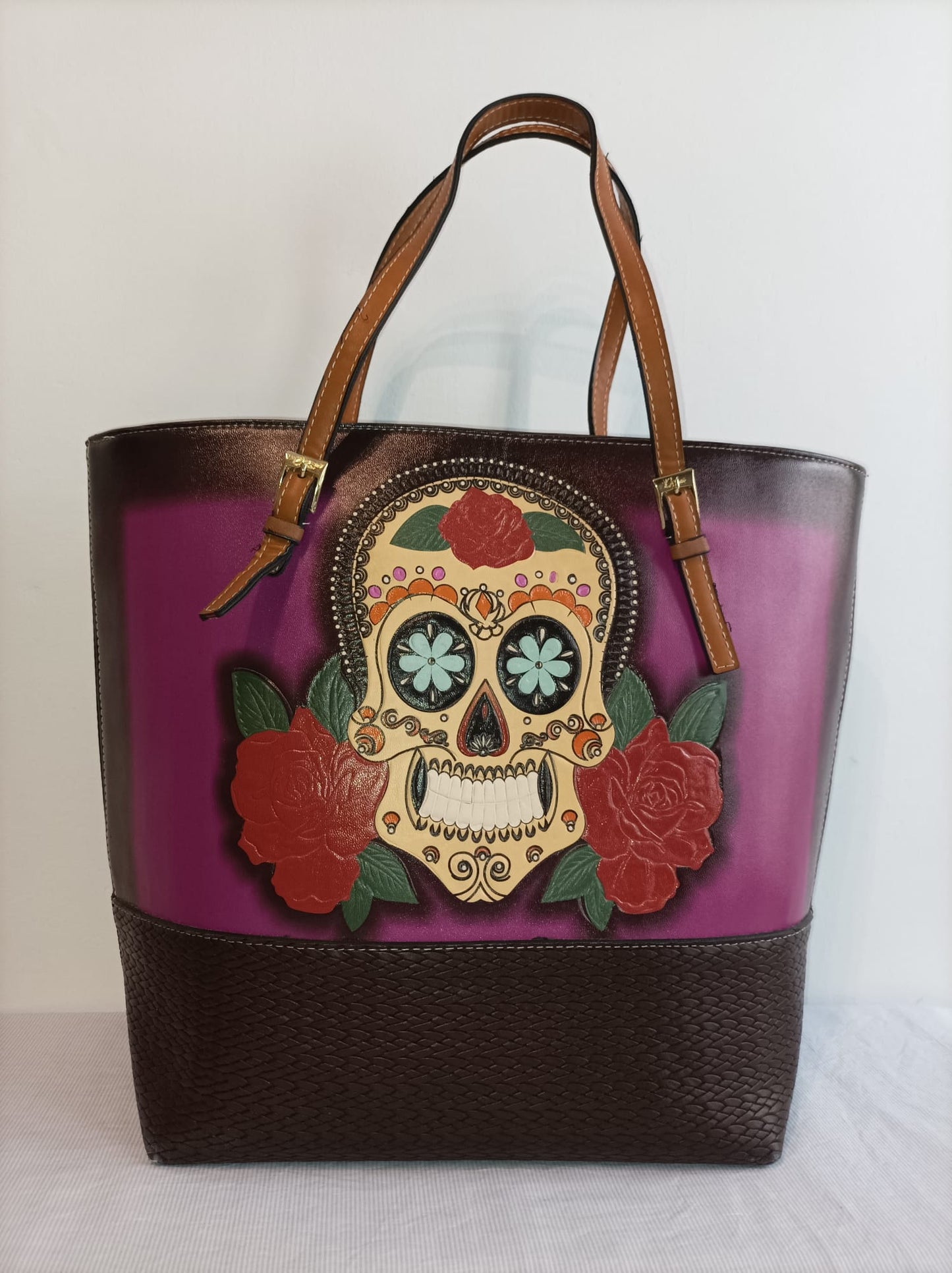 Bolso de mano con fondo morado, detalles en marrón y una calavera decorativa rodeada de rosas